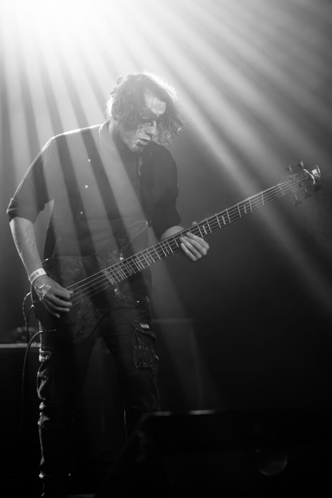 Baixista em concerto fotografado a preto e branco, atravessado por feixes de luz diagonais que destacam o músico e o instrumento.