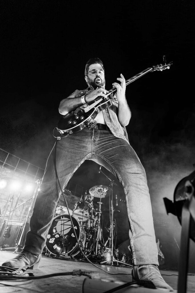 Guitarrista fotografado de baixo para cima durante um concerto, com perspetiva de grande angular e baterista em segundo plano.